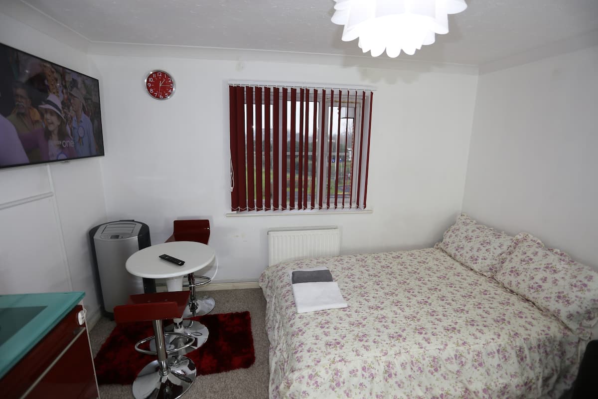A bedroom with a bed, a small table, a chair, a radiator, a clock, and a window with blinds.