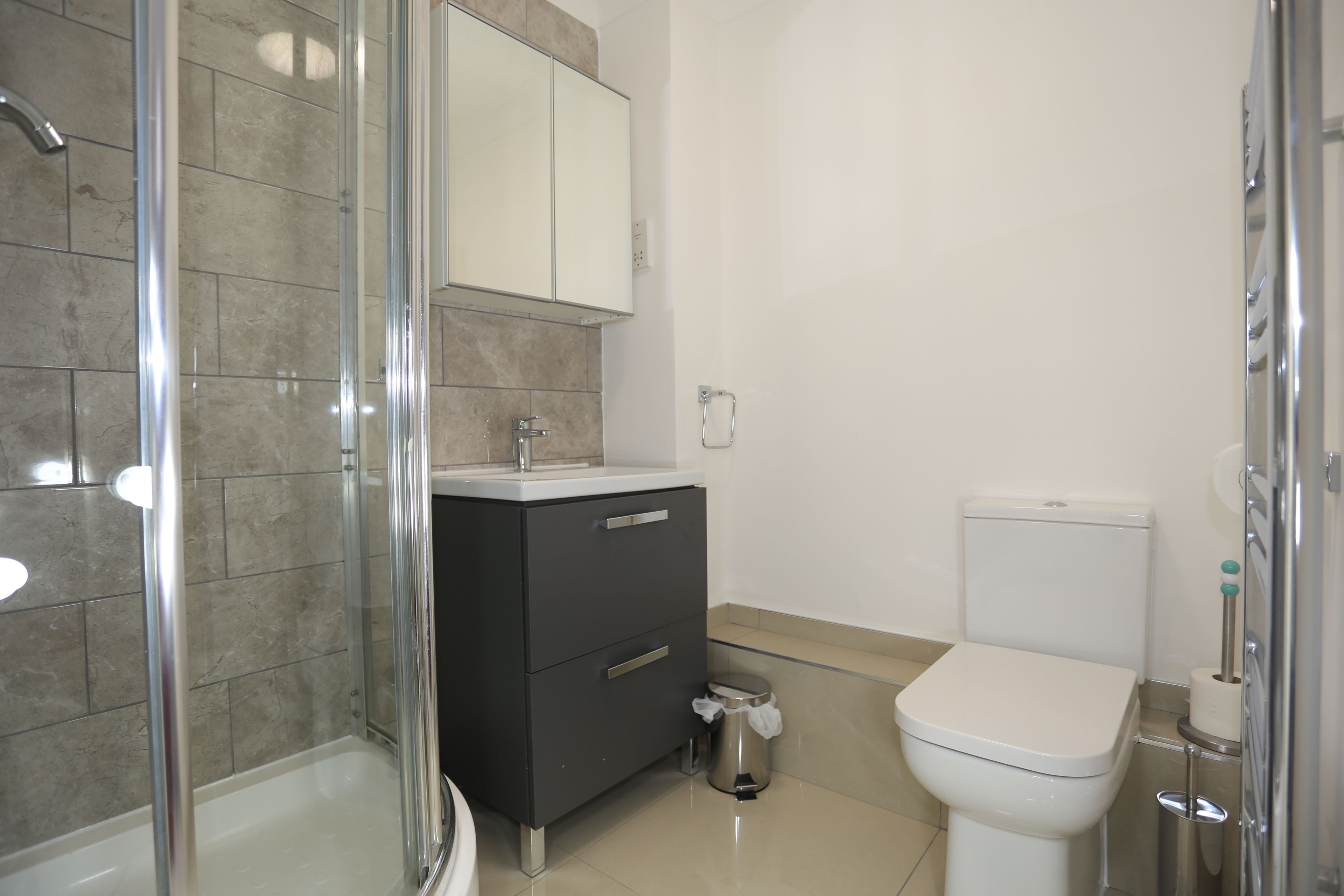 Et badeværelse med glasbrusekabine, et sort forfængelighedsbord med spejl, et hvidt toilet og en skraldespand.
