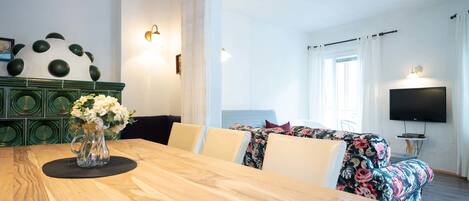 A modern living room with a wooden dining table, floral sofa, and a flat-screen TV mounted on the wall.