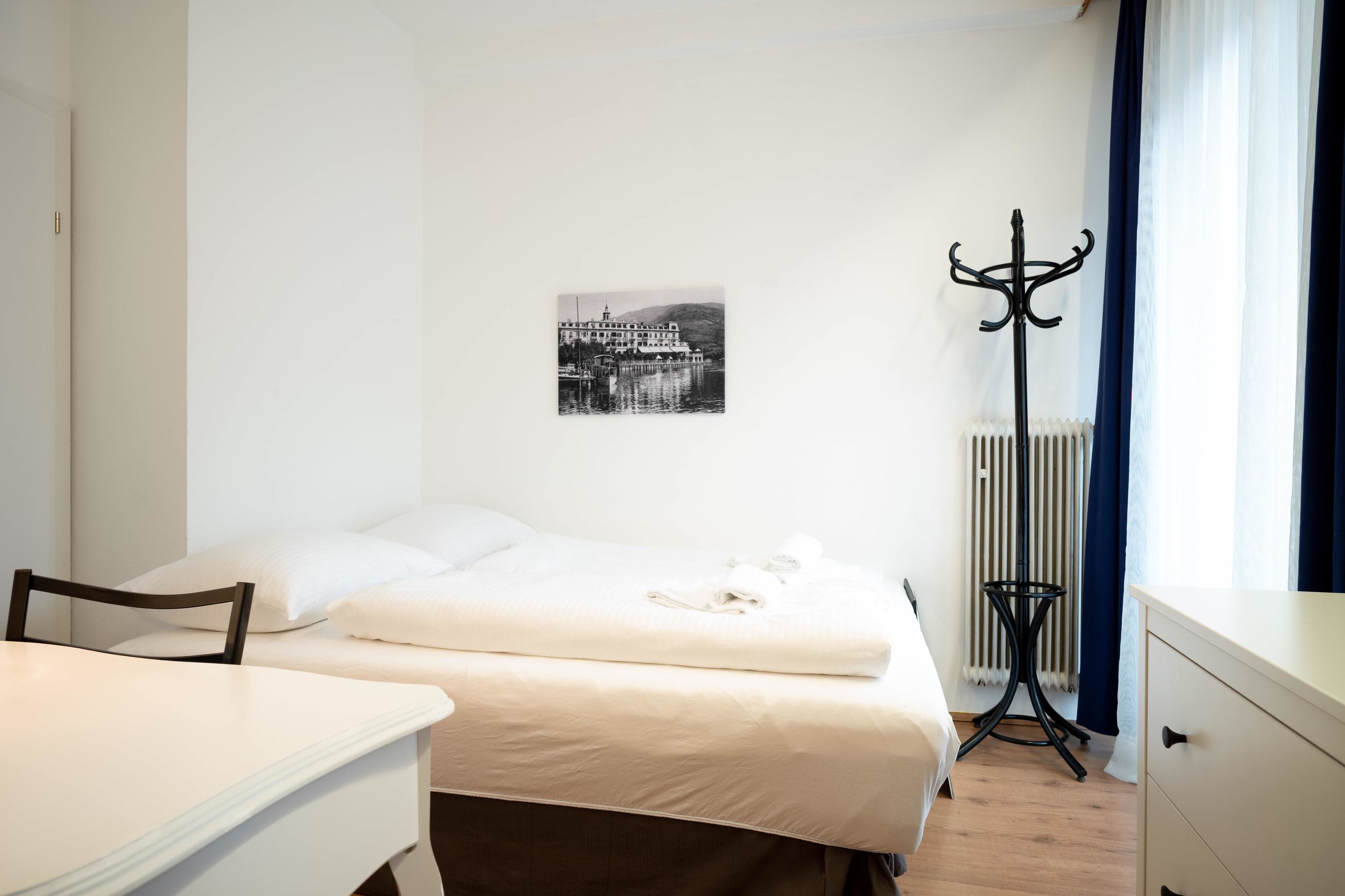 Un dormitorio con cama, una silla, una mesa, perchero y radiador.