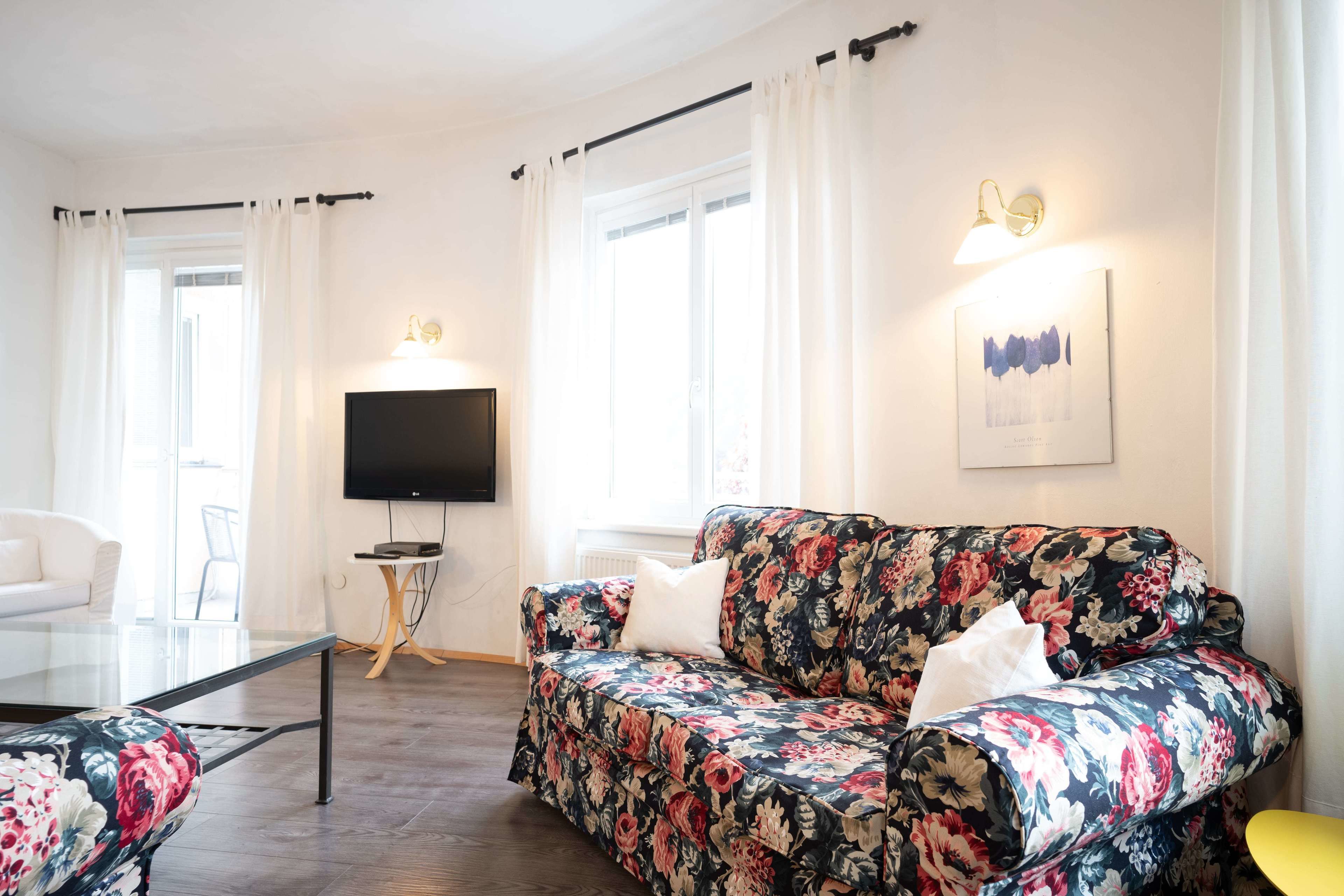 Ein Wohnzimmer mit einem blumigen Sofa, einem Glastisch, einem Flachbildfernseher und einem gerahmten Bild an der Wand.