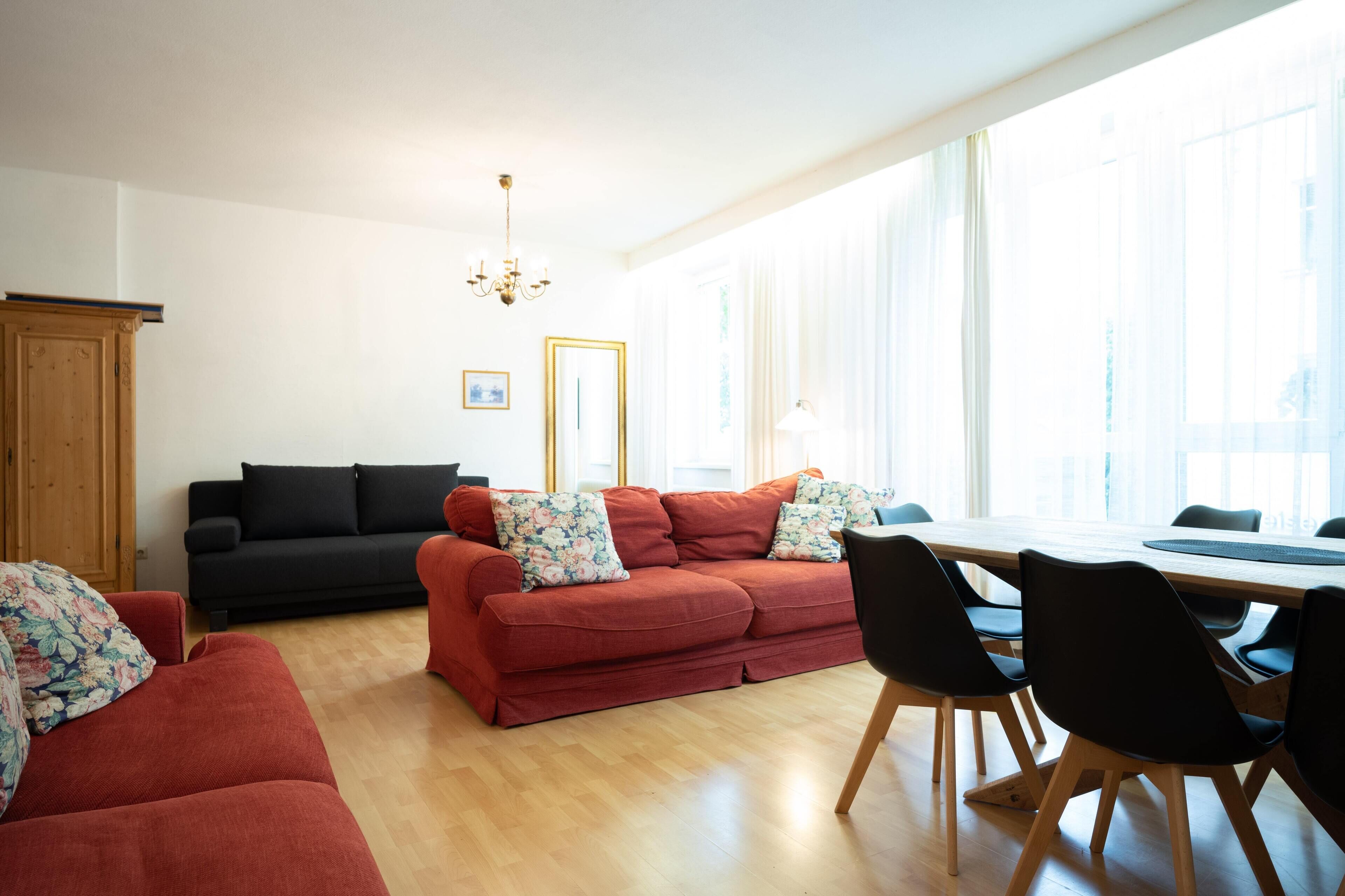 Ett modernt vardagsrum med en röd soffa, en svart soffa och ett matbord med stolar. Det finns en kristallkrona, ett träskåp och ett stort fönster med tunna, genomskinliga gardiner.