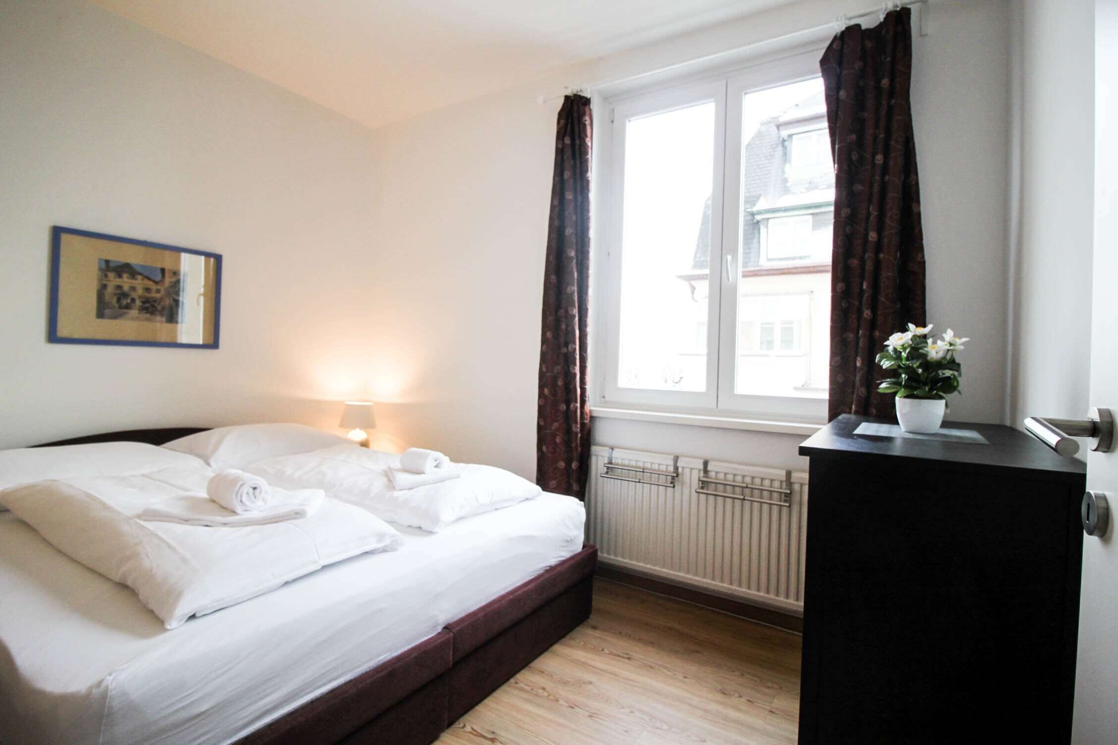Ein ordentlich bezogenes Bett mit weißen Leinen, eine Nachttischlampe, ein Heizkörper und ein Fenster mit Vorhängen.