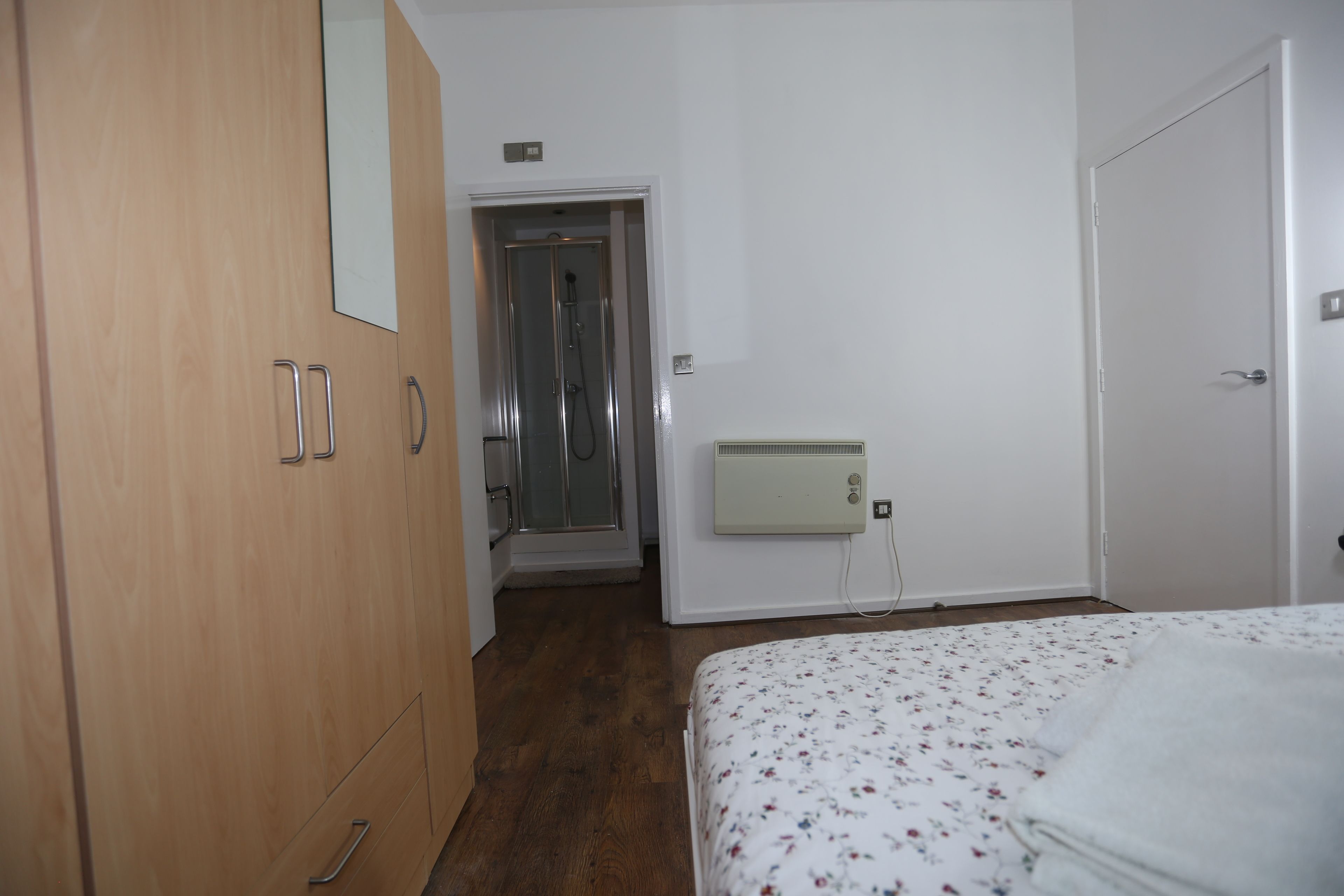 A bedroom with a wooden wardrobe, a bed with a floral duvet, a shower, and a radiator.