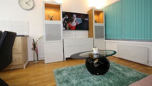 A modern living room with a glass coffee table, a clock on the wall, and a television.