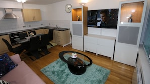 A modern kitchen with wooden cabinets, a dining area with a black table and chairs, and a living space with a sofa and a coffee table.