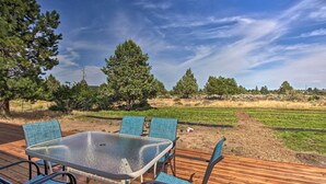 Una terraza al aire libre con una mesa de vidrio y sillas azules, rodeada de un campo de césped y árboles.