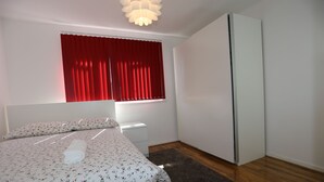 Ein Schlafzimmer mit Bett, Nachttisch, Kommode, einem Fenster mit roten Vorhängen und einem Kleiderschrank.