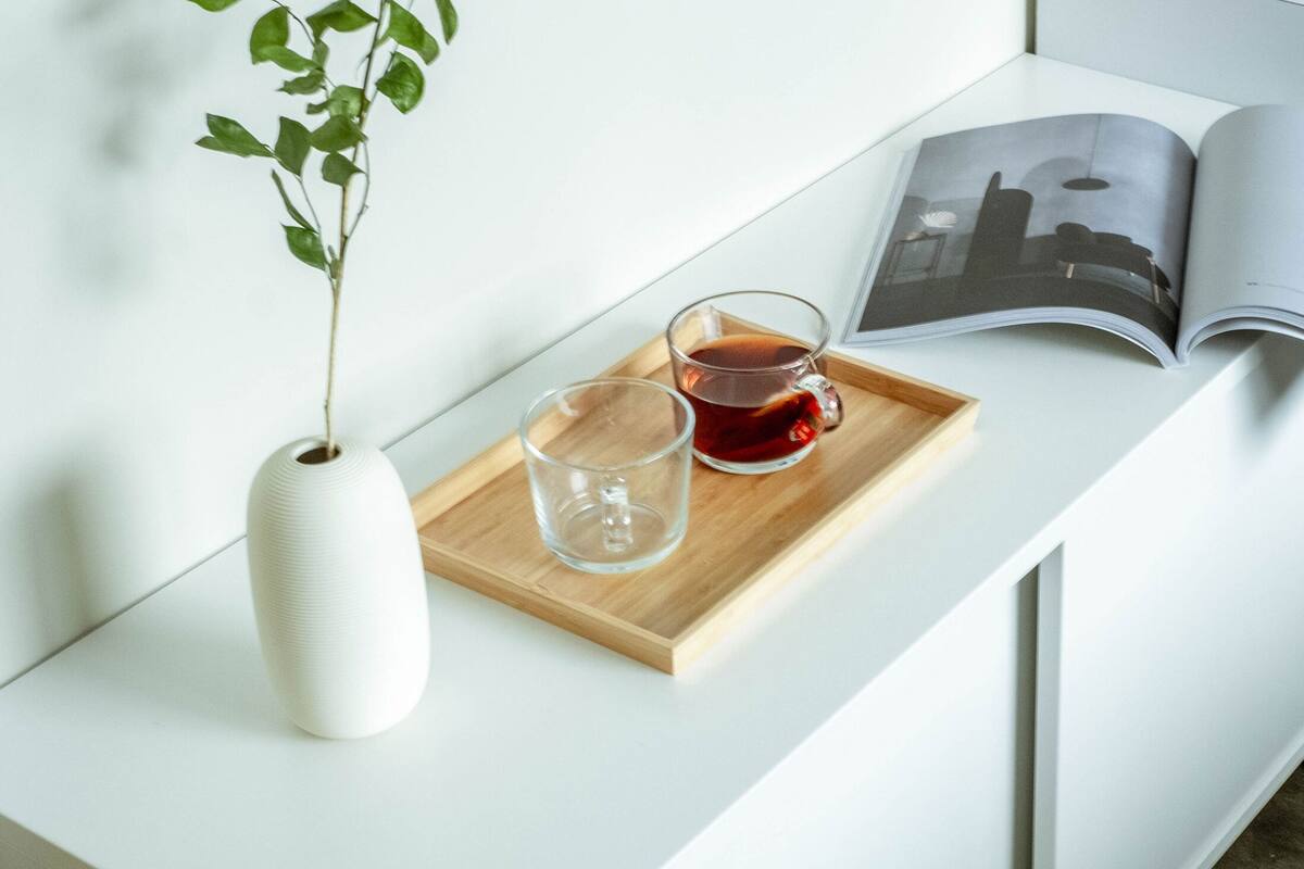 A white desk with a wooden tray holding a glass of tea and a clear glass. A white vase with green leaves is on the left. An open magazine is on the right.