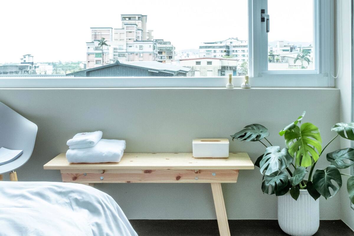 A hotel room with a bed, a desk with a chair, a window with a view of buildings, and a potted plant.