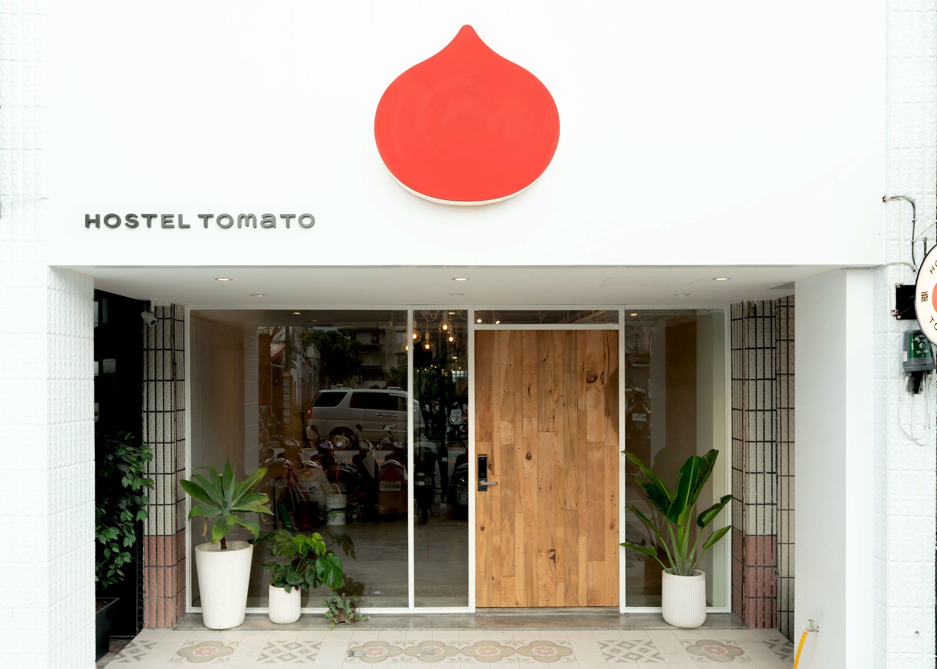 Entrada de um hostel com porta de madeira, plantas em vasos e um grande logotipo vermelho acima do nome 'HOSTEL TOMATO'.