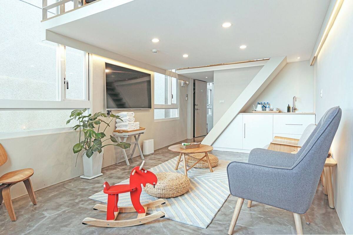 A modern living room with a high ceiling, a staircase, a kitchen area, and a large window providing natural light.