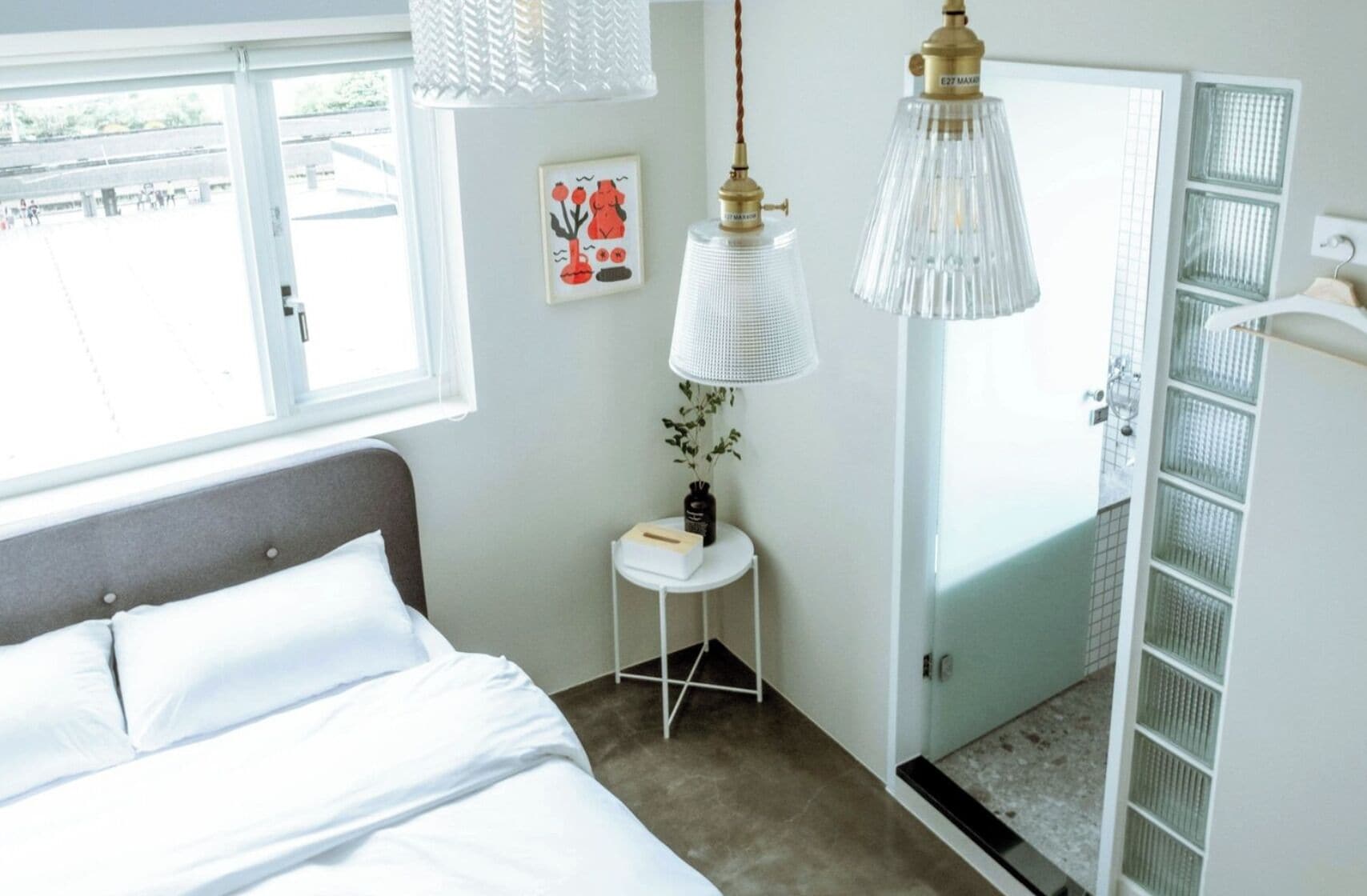 Un dormitorio con cama, una mesita de noche con lámpara, una ventana y una puerta que da al baño.