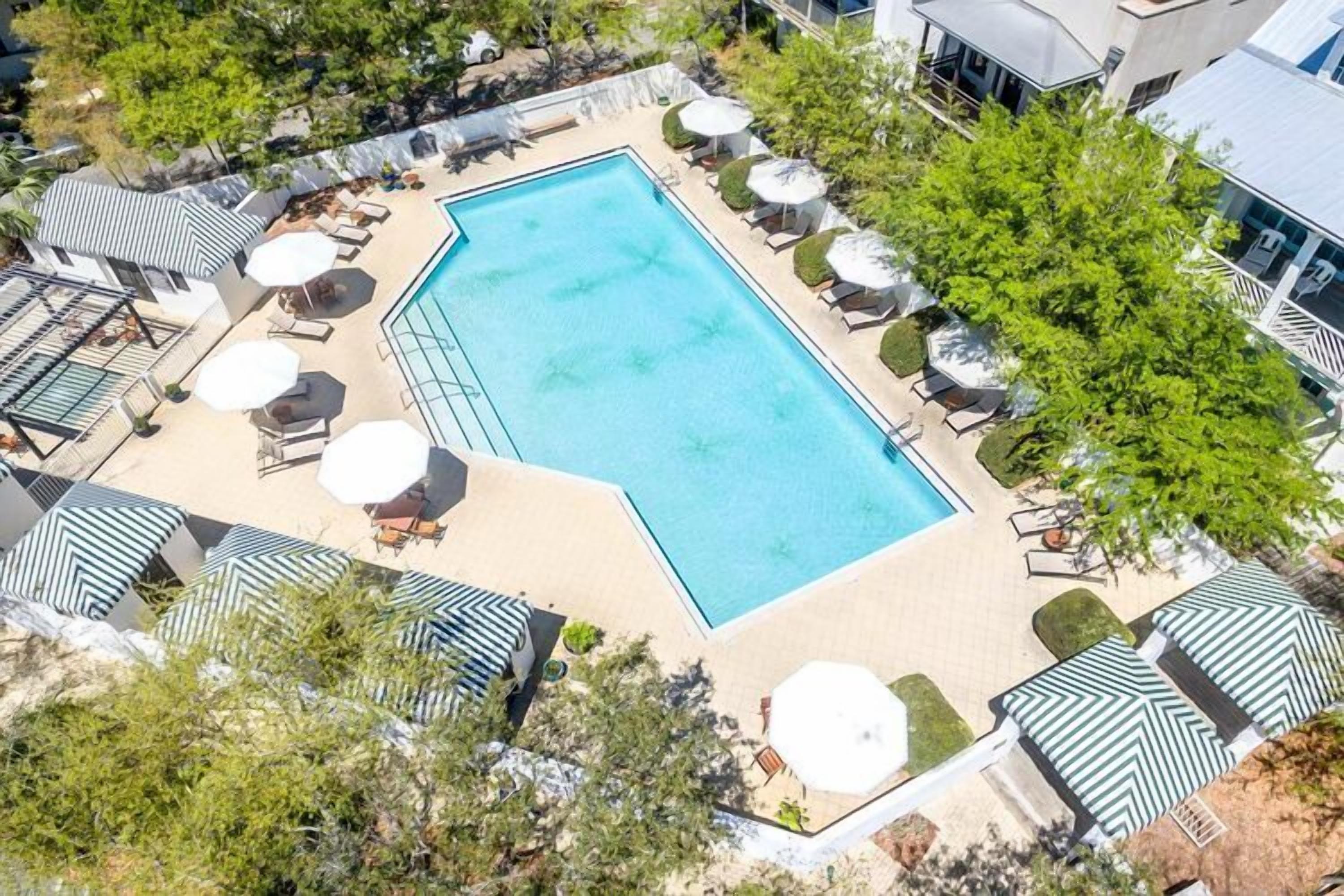 Vista aérea de una piscina rectangular rodeada de sillas de descanso y sombrillas, con árboles y edificios al fondo.