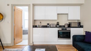 Eine kleine Küche mit weißen Schränken, einem eingebauten Backofen, einer Mikrowelle und einem Toaster. Eine Holztür führt in einen angrenzenden Raum.