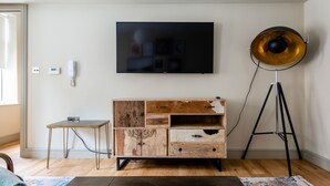Ein Zimmer mit einem Holzkasten, einem kleinen Tisch, einer Stehlampe und einem an der Wand befestigten Fernseher.