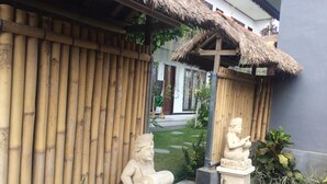 Una valla de bambú tradicional, una estructura con techo de paja y dos estatuas de piedra en un jardín.