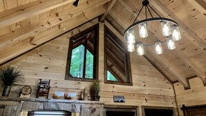 Interior de una cabaña de madera con techo inclinado, lámpara colgante, chimenea y ventana con vistas a la vegetación.
