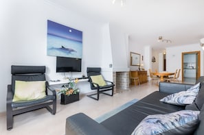 A modern living room with a sofa, two armchairs, a flat-screen TV, and a dining area with a table and chairs.