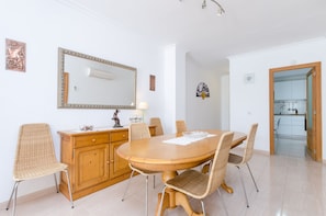 Un coin repas avec une table en bois, des chaises en osier, un buffet en bois, un grand miroir et une cuisine visible à travers une porte ouverte.