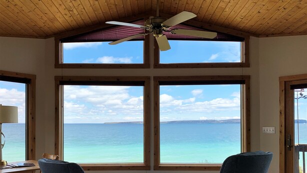 A room with a large window overlooking a body of water, a ceiling fan, and two blue chairs. - Huron Haven in St. Ignace (Saint Ignace)