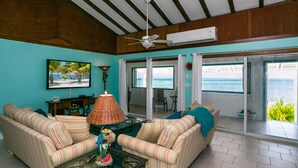 Ein Wohnzimmer mit gestreiften Sofas, einem Glastisch, einem Deckenventilator und einem großen Fernseher, der eine Strandansicht zeigt.