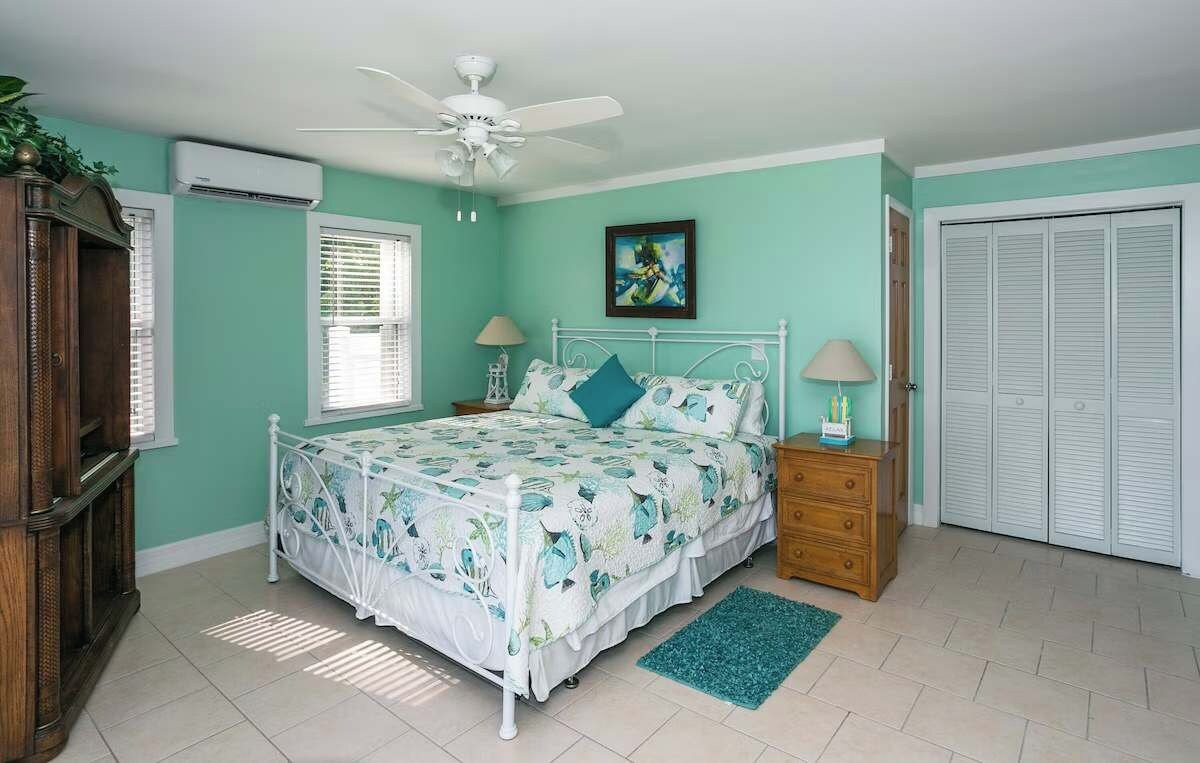 Un dormitorio con cama, mesitas de noche, ventilador de techo, aire acondicionado, ventana con persianas, puerta y alfombra verde.