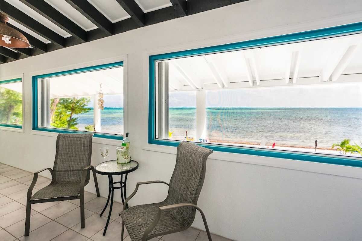 A room with two chairs and a small table, large windows with a view of the sea, and a ceiling with exposed beams.