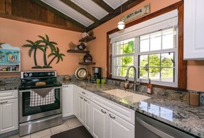 A kitchen with white cabinets, a granite countertop, a stove, and a window with a view of greenery. - The peace and tranquility of this cottage Works Wonders on the soul.... (Grand Cayman)