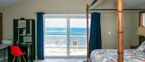 A bedroom with a bed, a desk, a chair, and a view of the sea through a balcony.