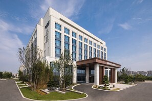 A modern hotel building with a prominent entrance, large windows, and a well-maintained landscaped area with trees and shrubs.