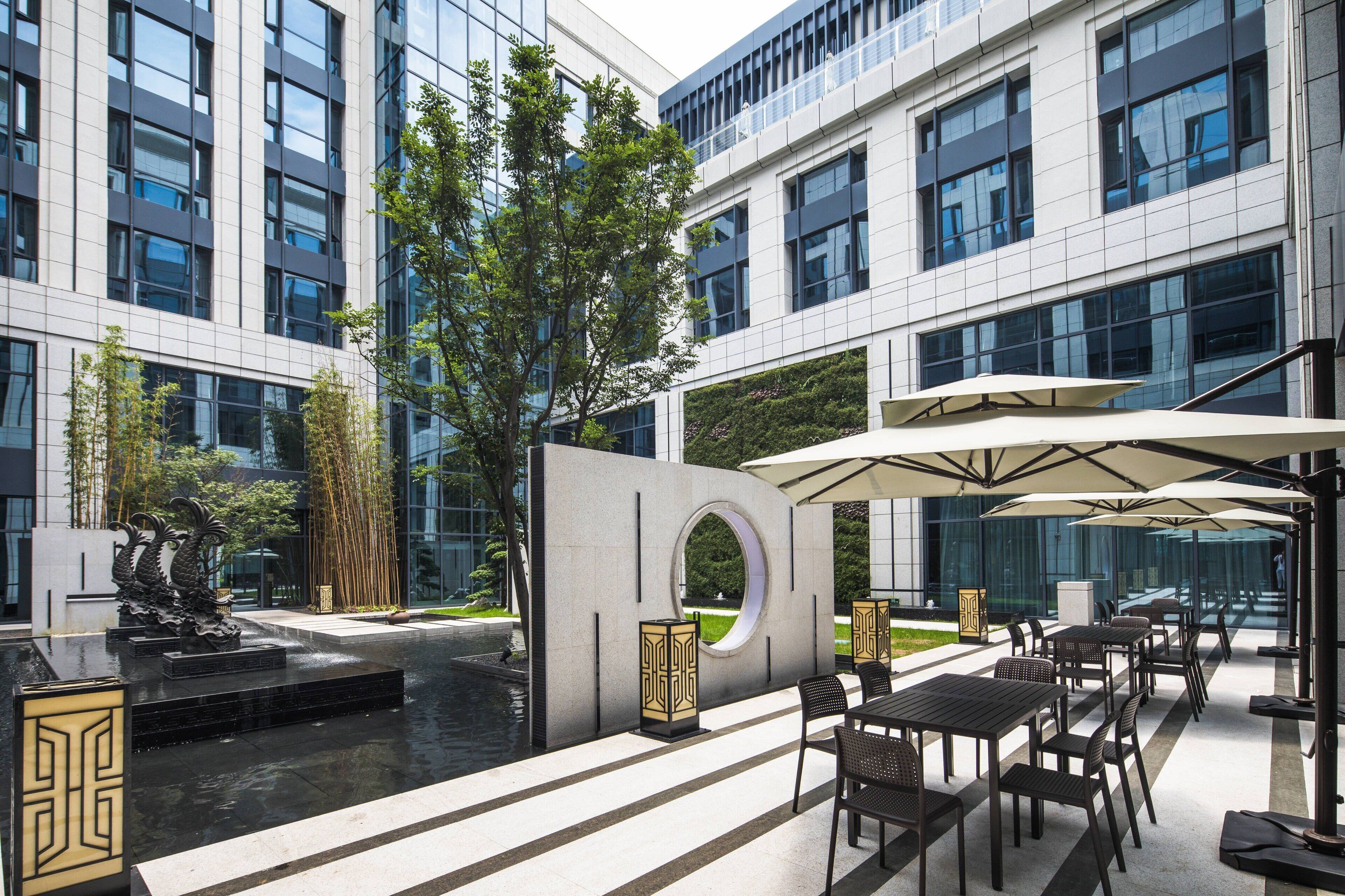 An outdoor seating area with tables and umbrellas, a fountain, and a modern building with large windows.
