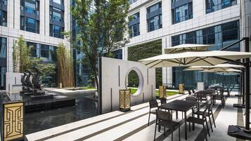 An outdoor seating area with tables and umbrellas, a fountain, and a modern building with large windows.