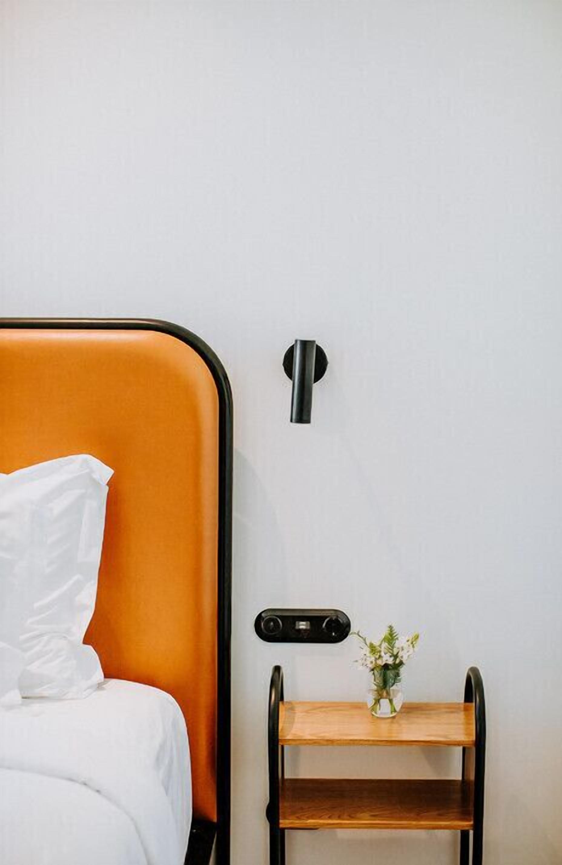 A bed with white bedding and an orange headboard, a small wooden nightstand with a vase of flowers, and a wall-mounted control panel.