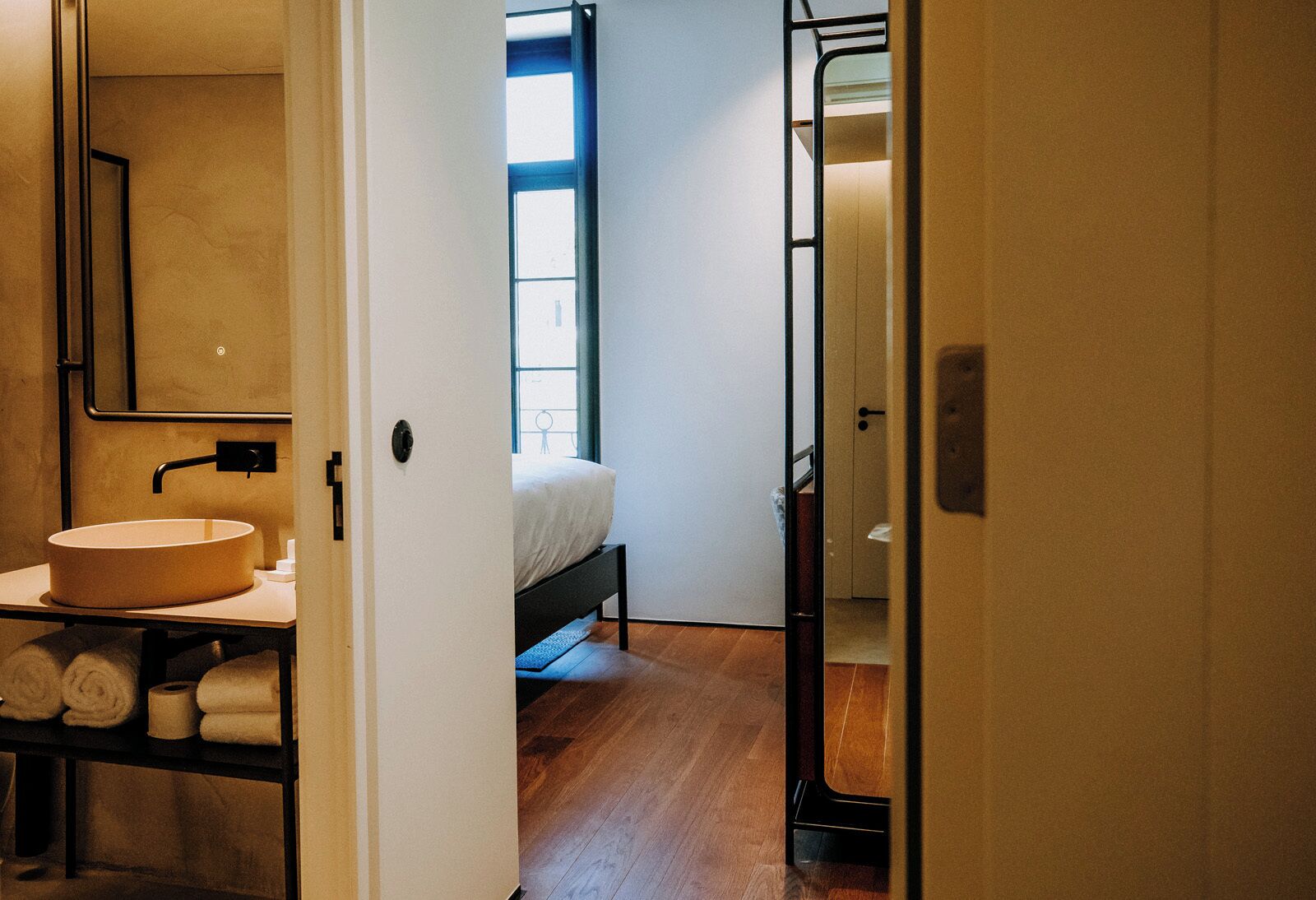 A modern bathroom with a round sink, a mirror, and a toilet. There is a bed with a white mattress and a window above it.