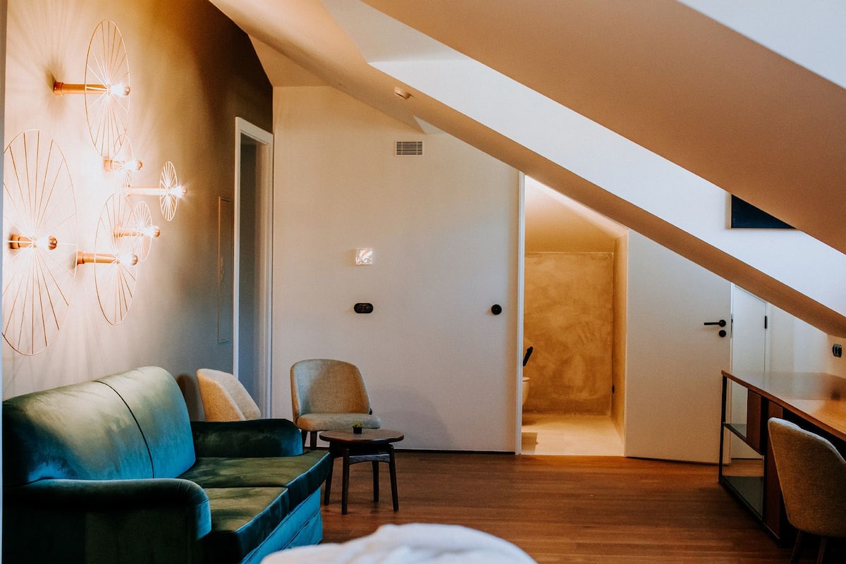 A modern loft bedroom with a sloped ceiling, a green sofa, a chair, and a dining table.