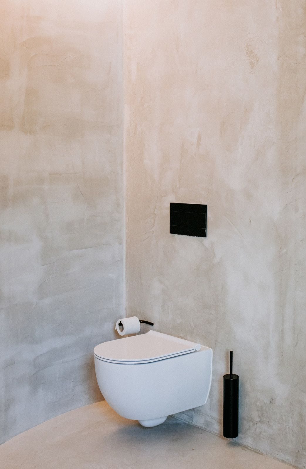 A minimalist bathroom with a white toilet, a black flush tank, and a roll of toilet paper on the wall.