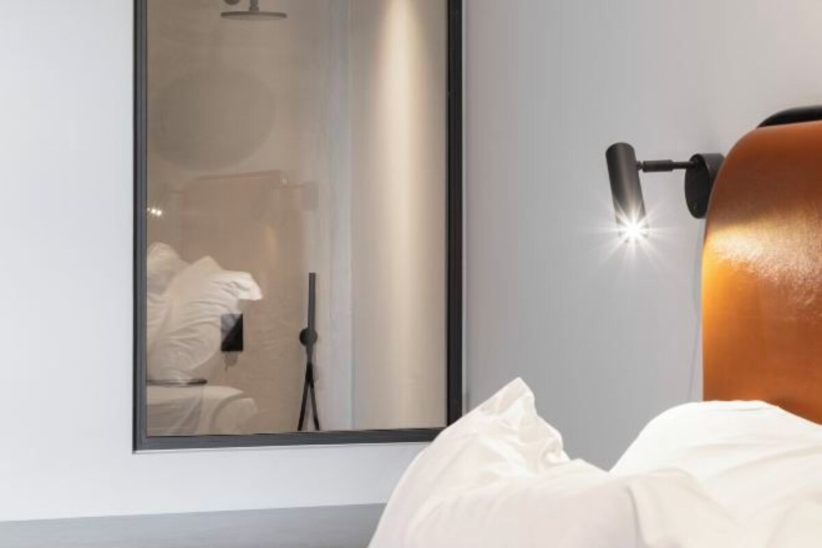 A bedroom with a bed, a wall-mounted lamp, and a mirror reflecting a bathroom.