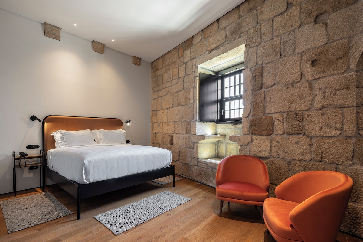 A bedroom with a stone wall, a bed, an orange chair, and a window with bars.