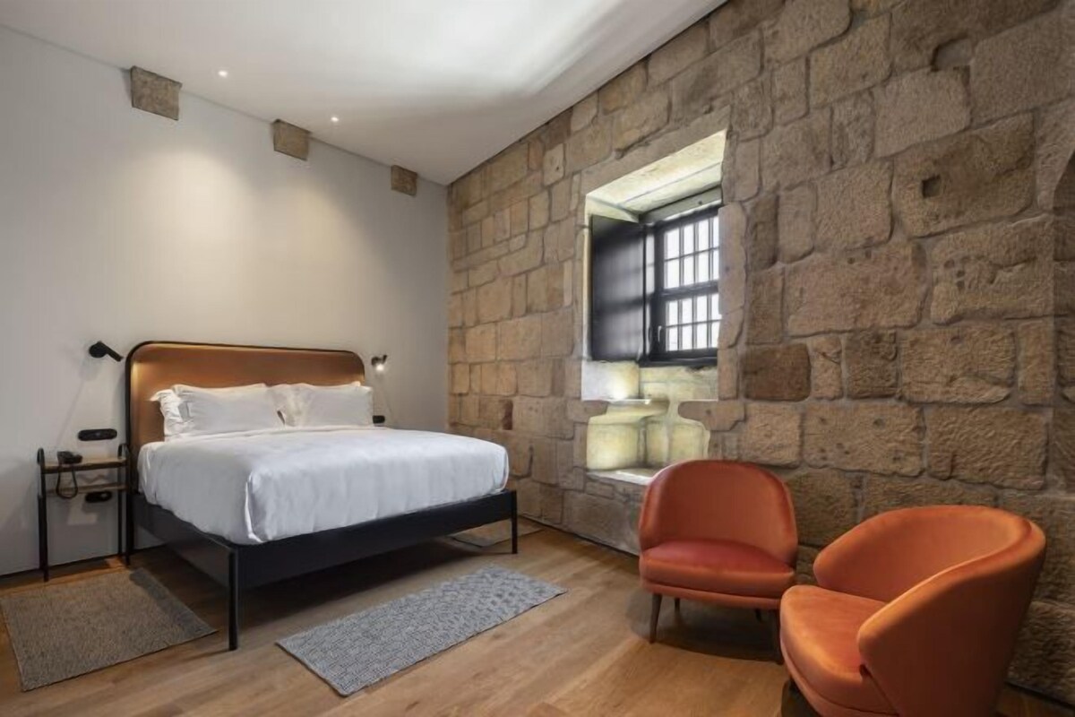 A bedroom with a stone wall, a bed, a nightstand, a chair, and a window.
