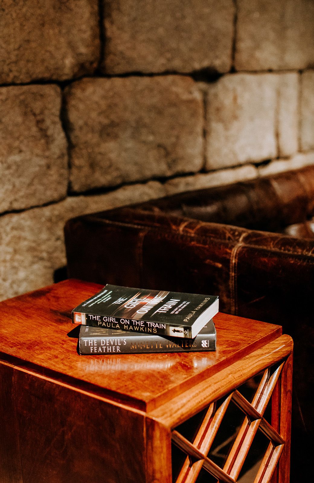 A stack of books on a wooden table with a stone wall in the background.