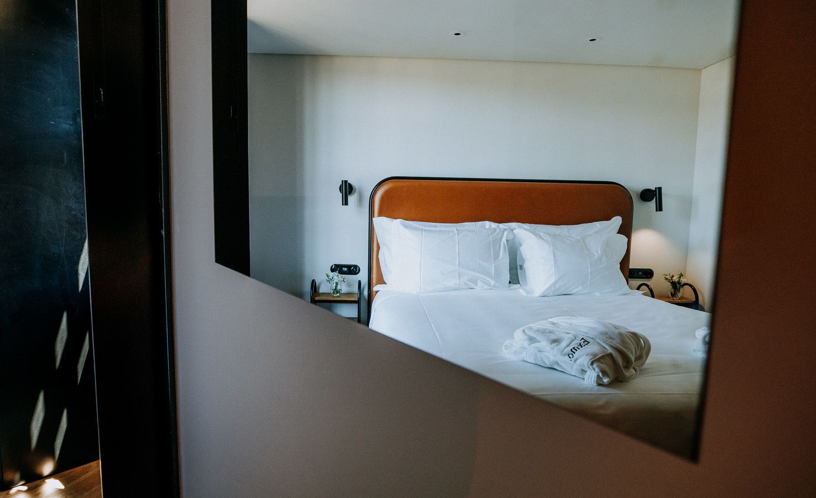 A hotel room with a bed, white linens, a wooden headboard, bedside tables, and a nightstand with a lamp.