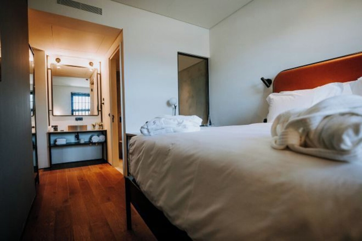A hotel room with a wooden floor, a bed with white bedding, a mirror, and a small desk with a lamp.
