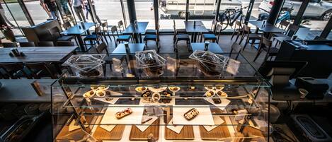 A bakery display with various pastries and desserts in a glass case, situated in a cafe with tables and chairs.