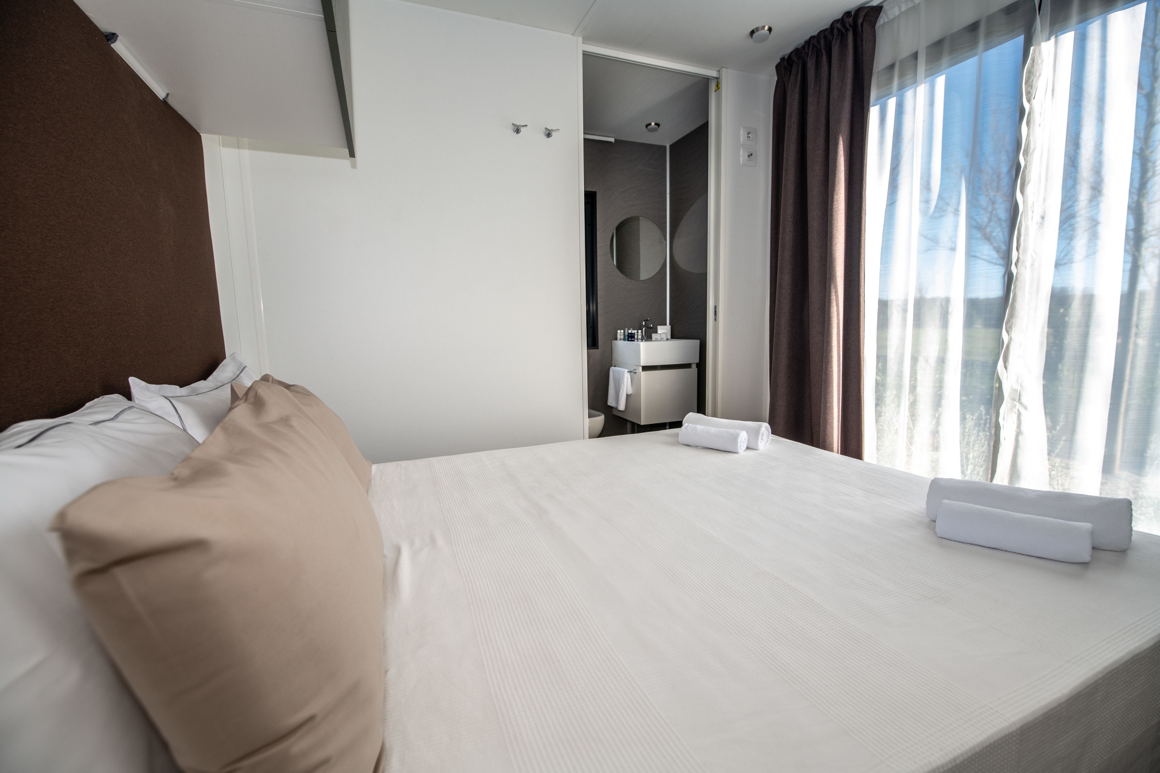 A hotel room with a large bed, a bathroom with a round mirror, and a view of trees through the window.