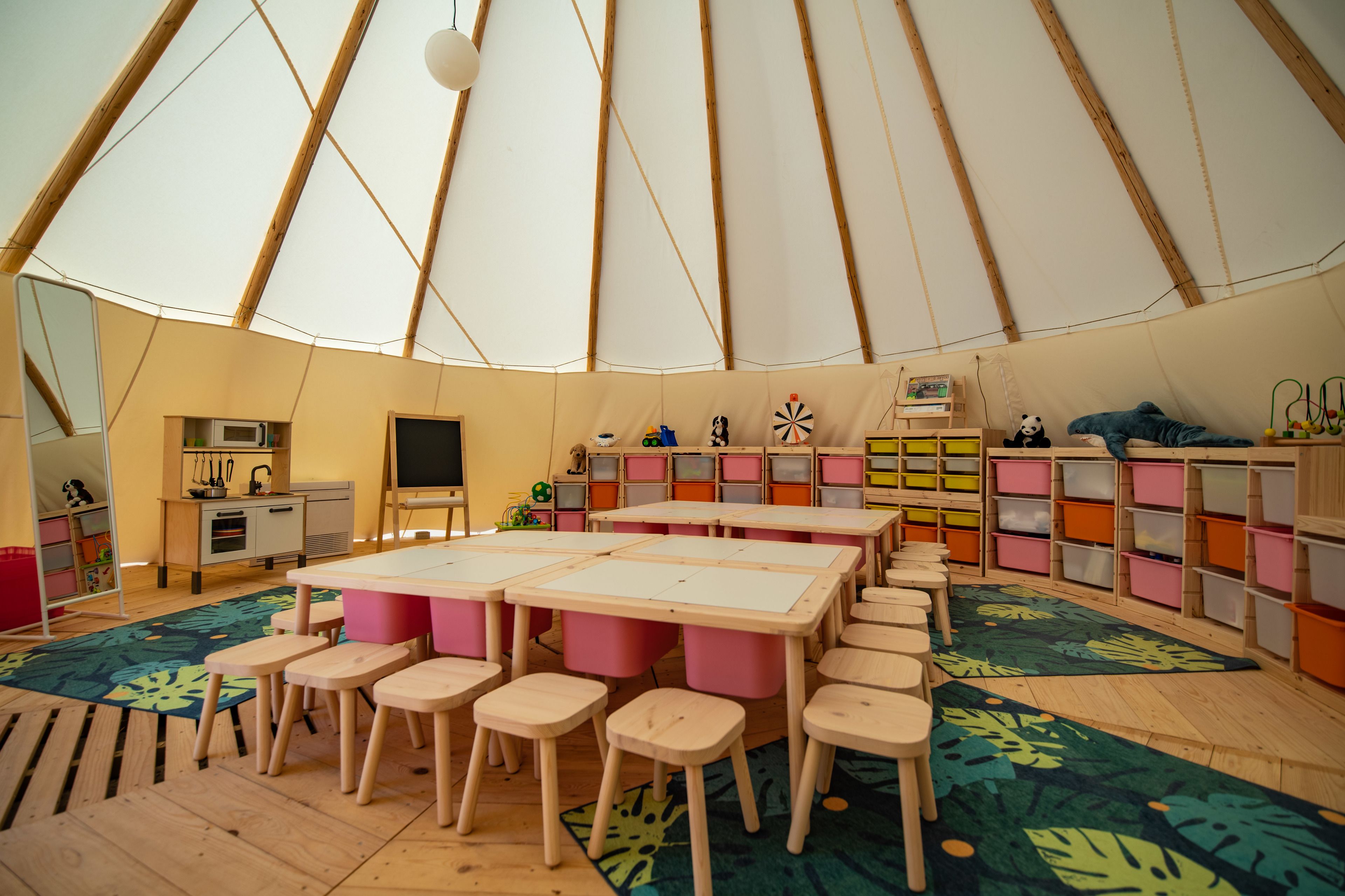 Un espacio interior amplio con techo alto y zona diáfana. Hay varias mesas y sillas dispuestas para actividades grupales. La sala cuenta con varios almacenajes y un televisor.