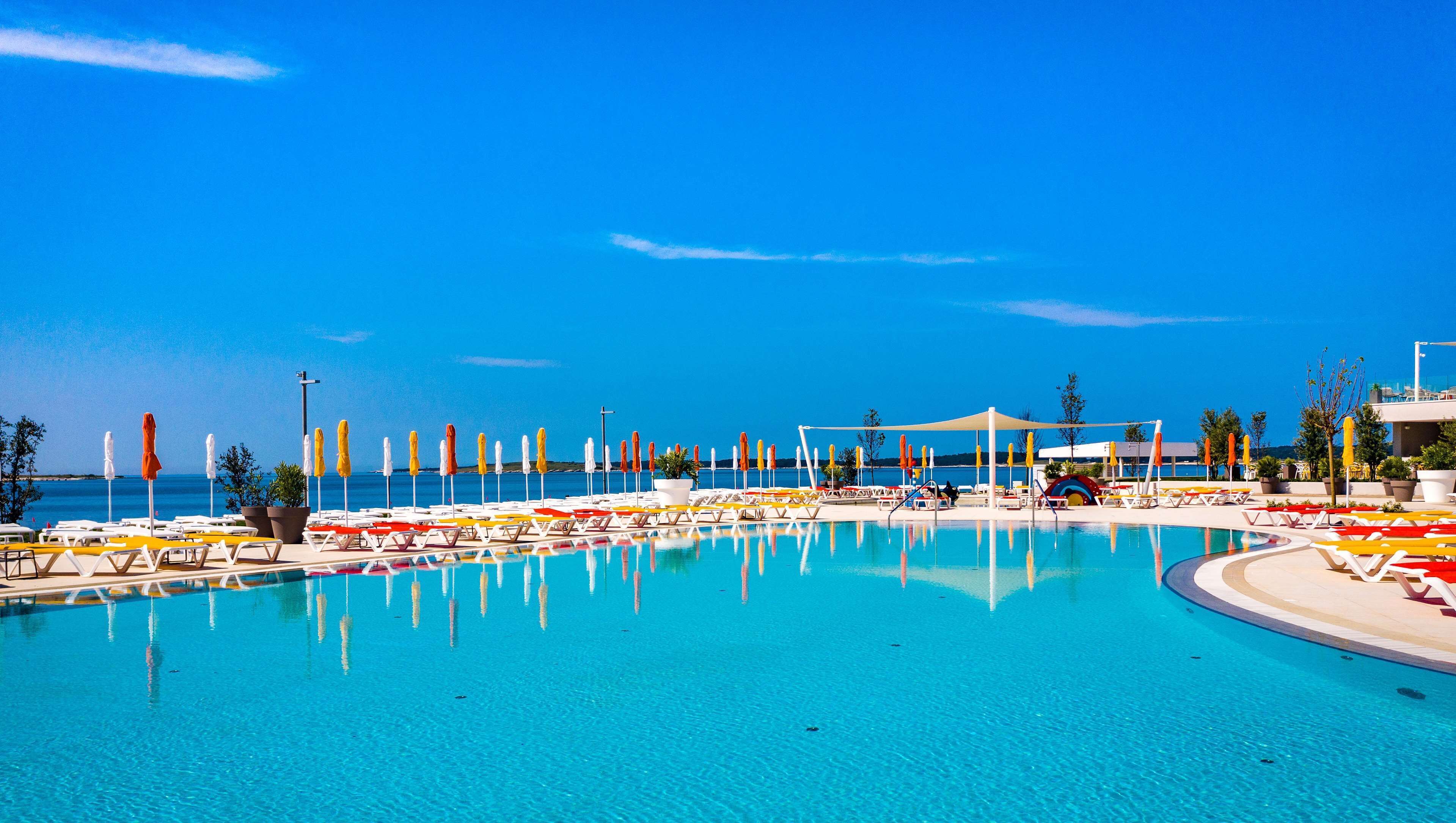 Ein Pool in einer Ferienanlage mit klarem, blauem Wasser, umgeben von Liegestühlen und Sonnenschirmen, mit Blick auf das Meer und die Hügel im Hintergrund.