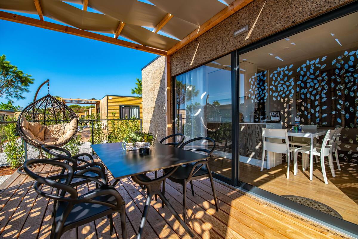 An outdoor patio with a wooden table and chairs, a hanging chair, and a glass door leading to an interior dining area.