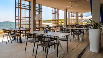 A spacious outdoor dining area with multiple tables and chairs, large windows offering a view of the sea, and a ceiling fan.