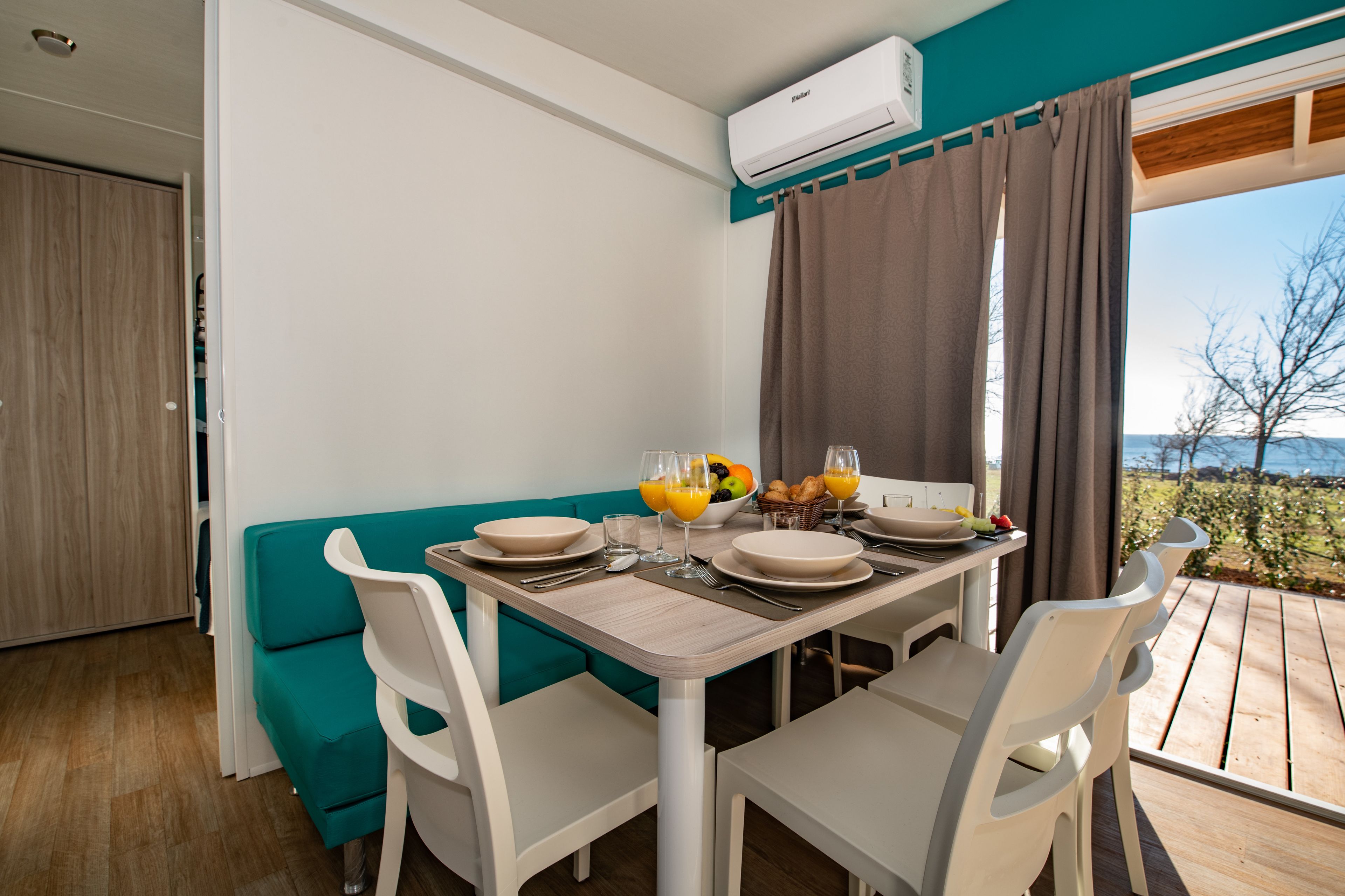 Un comedor con una mesa dispuesta para cuatro personas, un sofá color esmeralda, sillas blancas y vistas al exterior.